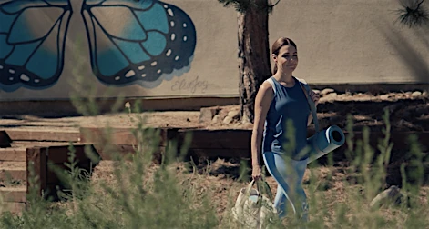 Woman carrying a yoga mat and bag walking outdoors past a wall with painted blue butterfly wings.