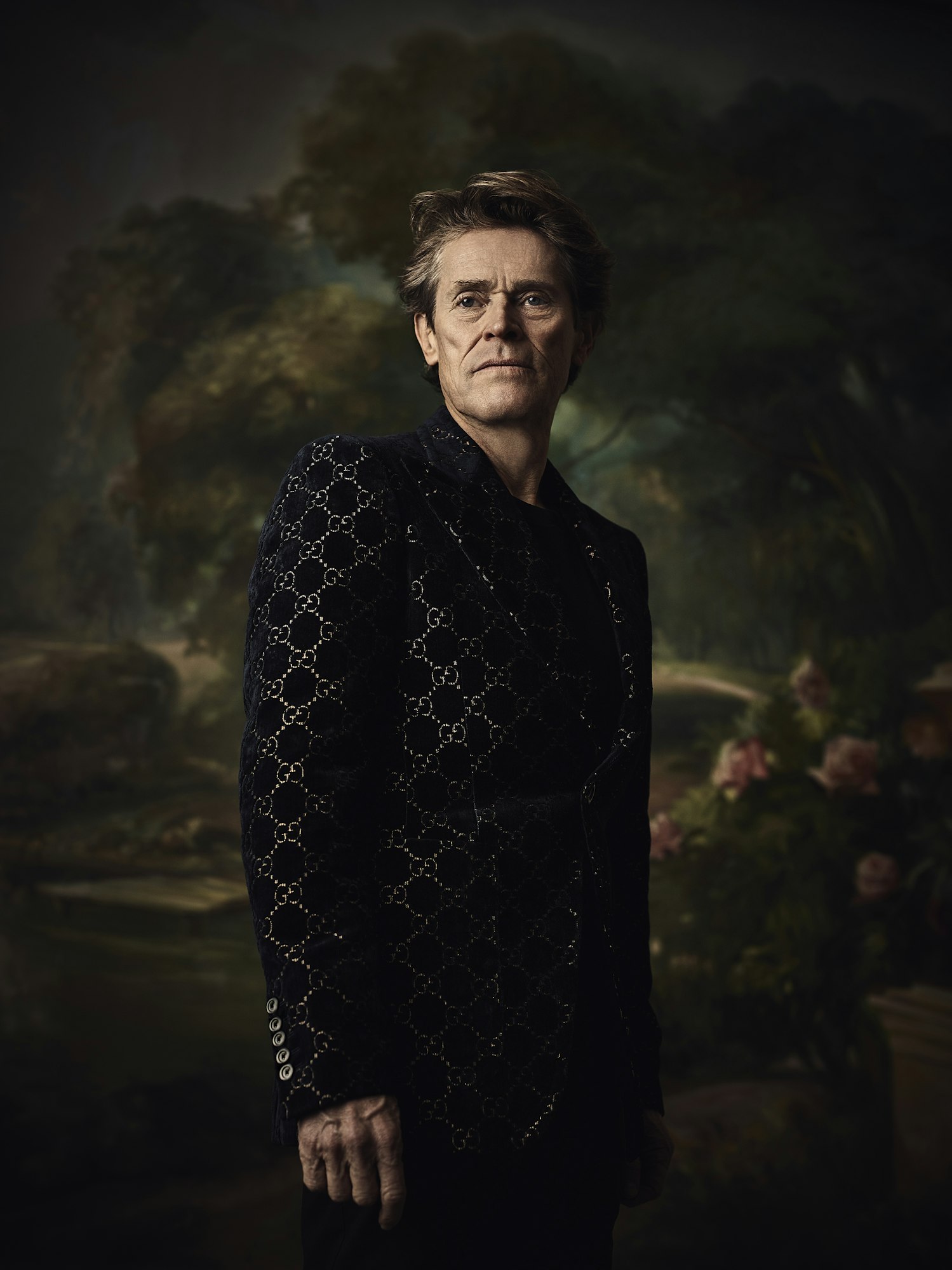 Portrait of older adult male in patterned jacket, posing indoors against a floral backdrop in a classic, formal style.