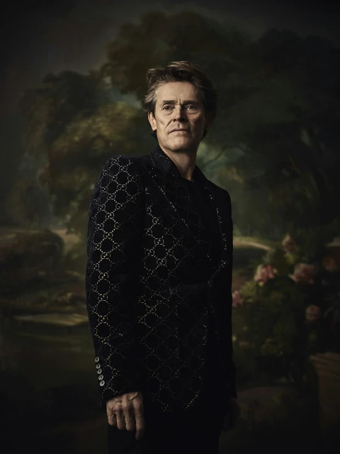 Portrait of older adult male in patterned jacket, posing indoors against a floral backdrop in a classic, formal style.