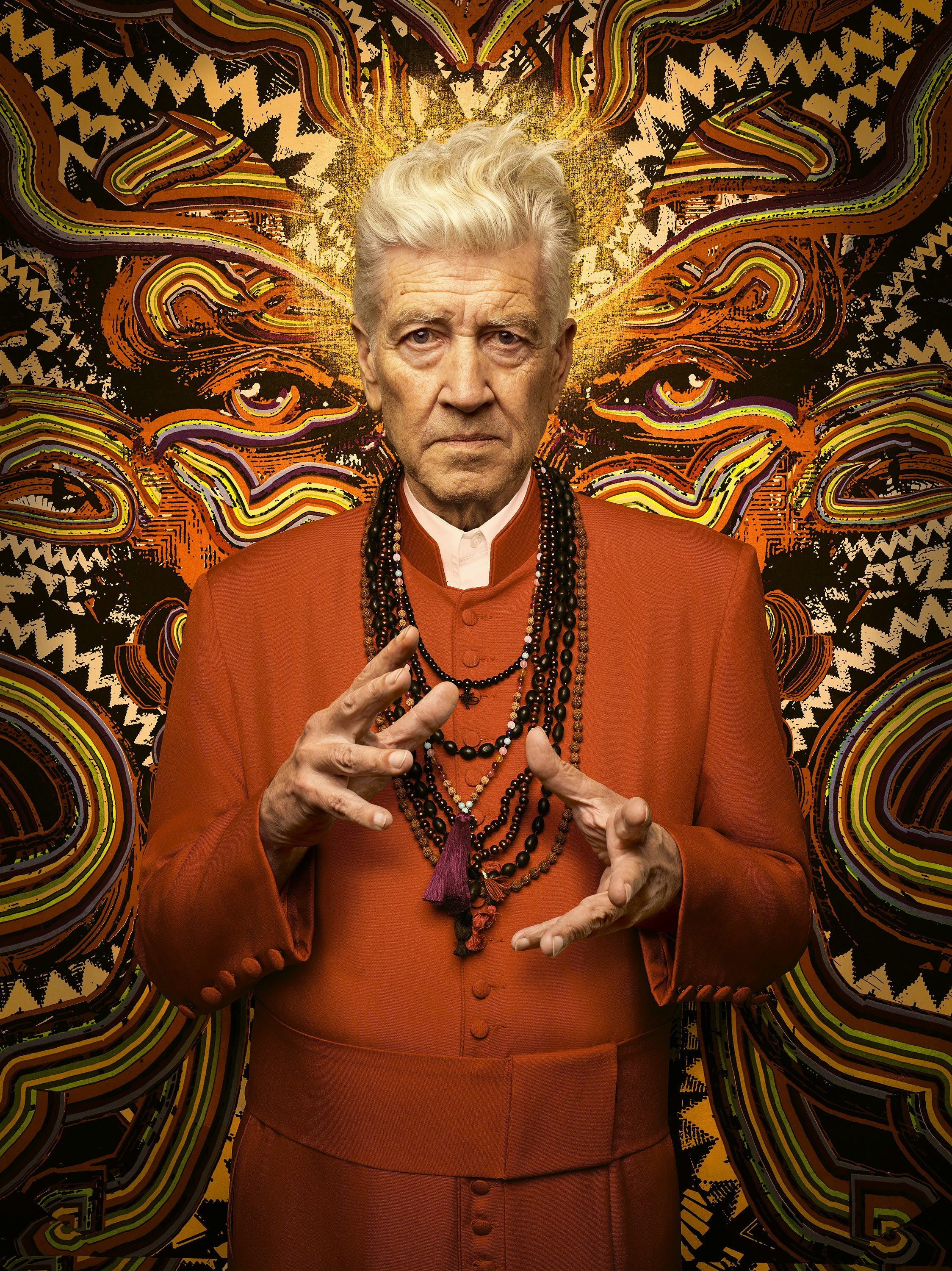 Portrait of David Lynch with orange robe and layered beaded necklaces, against a patterned backdrop with stylized eyes and sunburst.