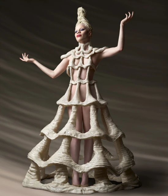 Albino woman wearing a cream-colored rope dress and headdress against a mottled gray background with arms outstretched, eye-level angle.