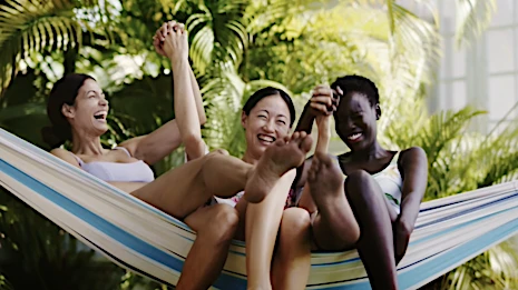 Three women in swimsuits laugh together while lying in a hammock surrounded by tropical greenery on a bright day.