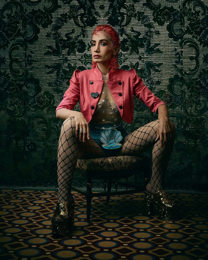 Adult female with pink hair and jacket sitting on a stool in front of a damask background, wearing fishnets and platform shoes.