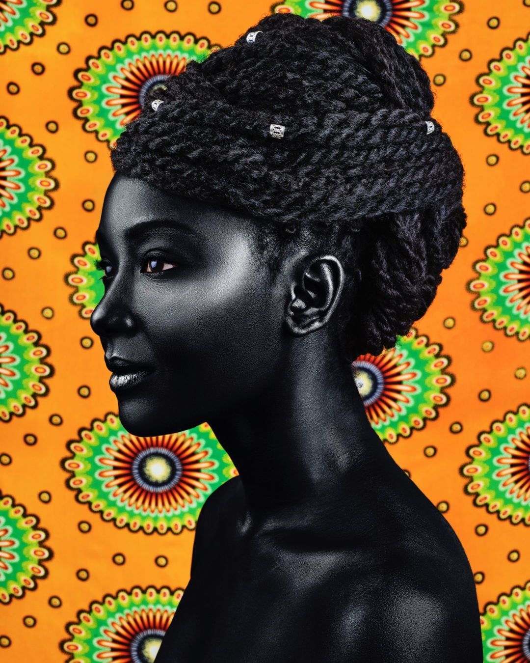 Profile portrait of a Black woman with an intricate updo against a vibrant patterned background. Features rich, dark skin and elaborate hair.