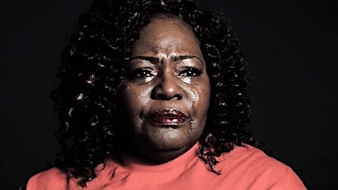 Close-up portrait of a crying African-American woman with dark curly hair wearing an orange shirt against a dark background.