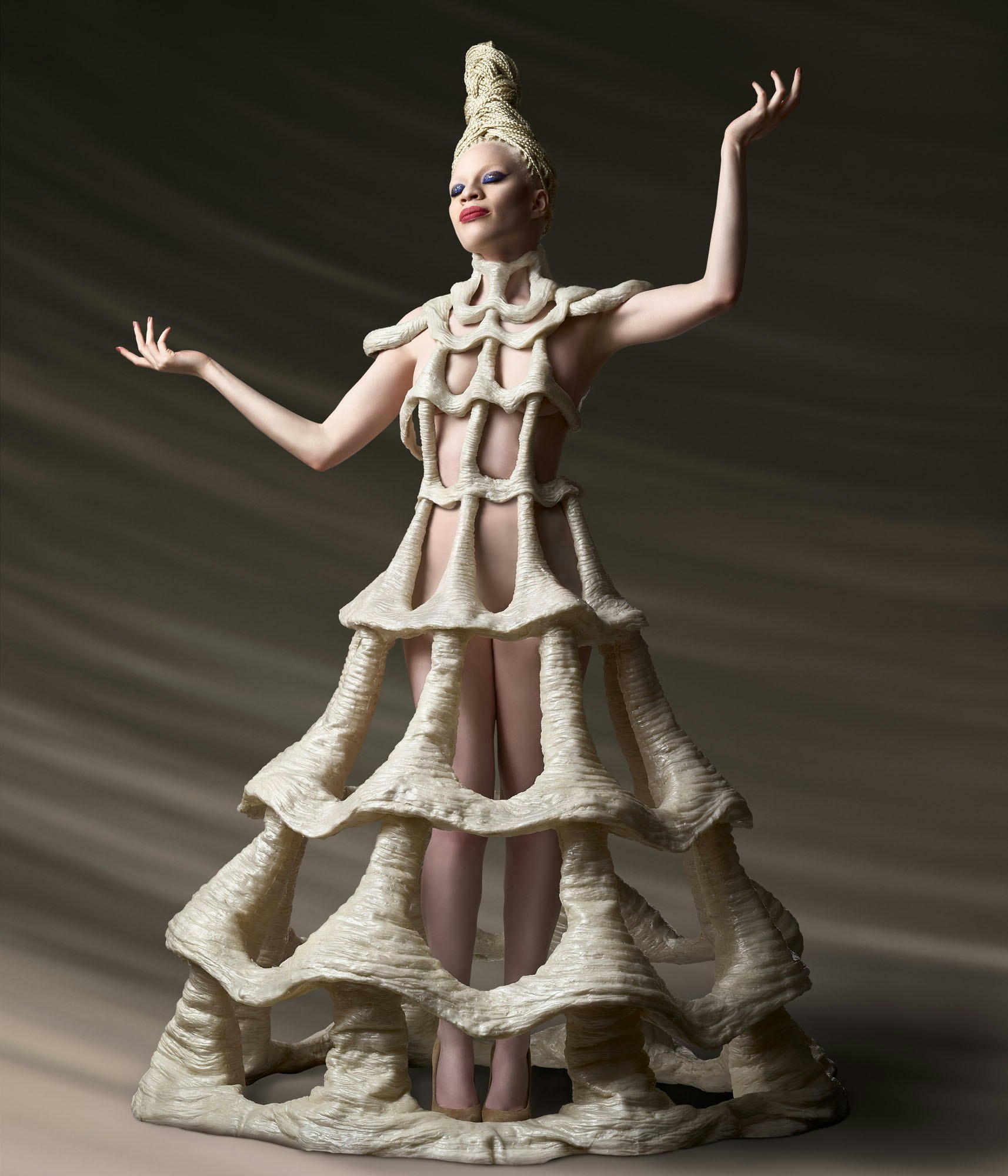 Albino model in unique sculptural dress posing in photo studio with outstretched arms and hands, dramatic lighting and earth-tone backdrop.