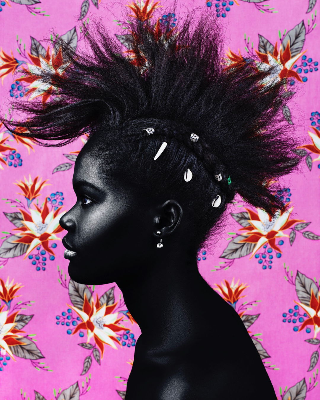 Profile portrait of a Black woman with a punk-style mohawk and shell hair accessories, set against a pink floral patterned background.