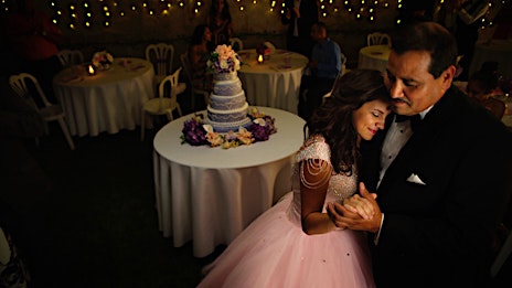 Bride and father dancing at wedding reception with cake table and guests. The event is lit by string lights.
