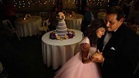 Bride and father dancing at wedding reception with cake table and guests. The event is lit by string lights.