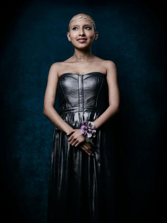Portrait of a smiling bald adult woman with a metallic dress and floral wristlet in front of a textured dark blue background.
