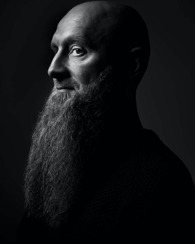 Monochrome portrait of a bald adult male with a long beard, looking to the side, in a studio with dark lighting and neutral background.
