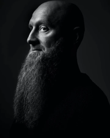 Monochrome portrait of a bald adult male with a long beard, looking to the side, in a studio with dark lighting and neutral background.