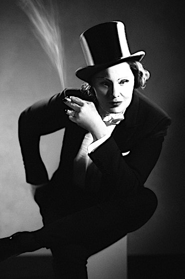 Black and white portrait of a person in a tuxedo and top hat holding a cigarette with smoke rising, photographed by Sandro.