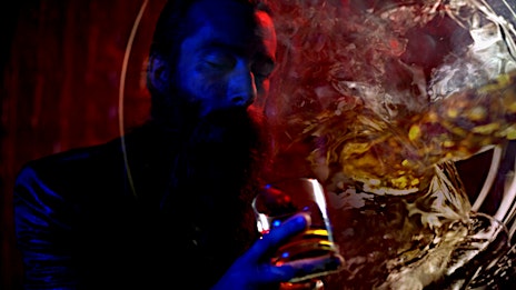 Man with beard holding a glass of amber liquid with moody red and blue lighting by Sandro.