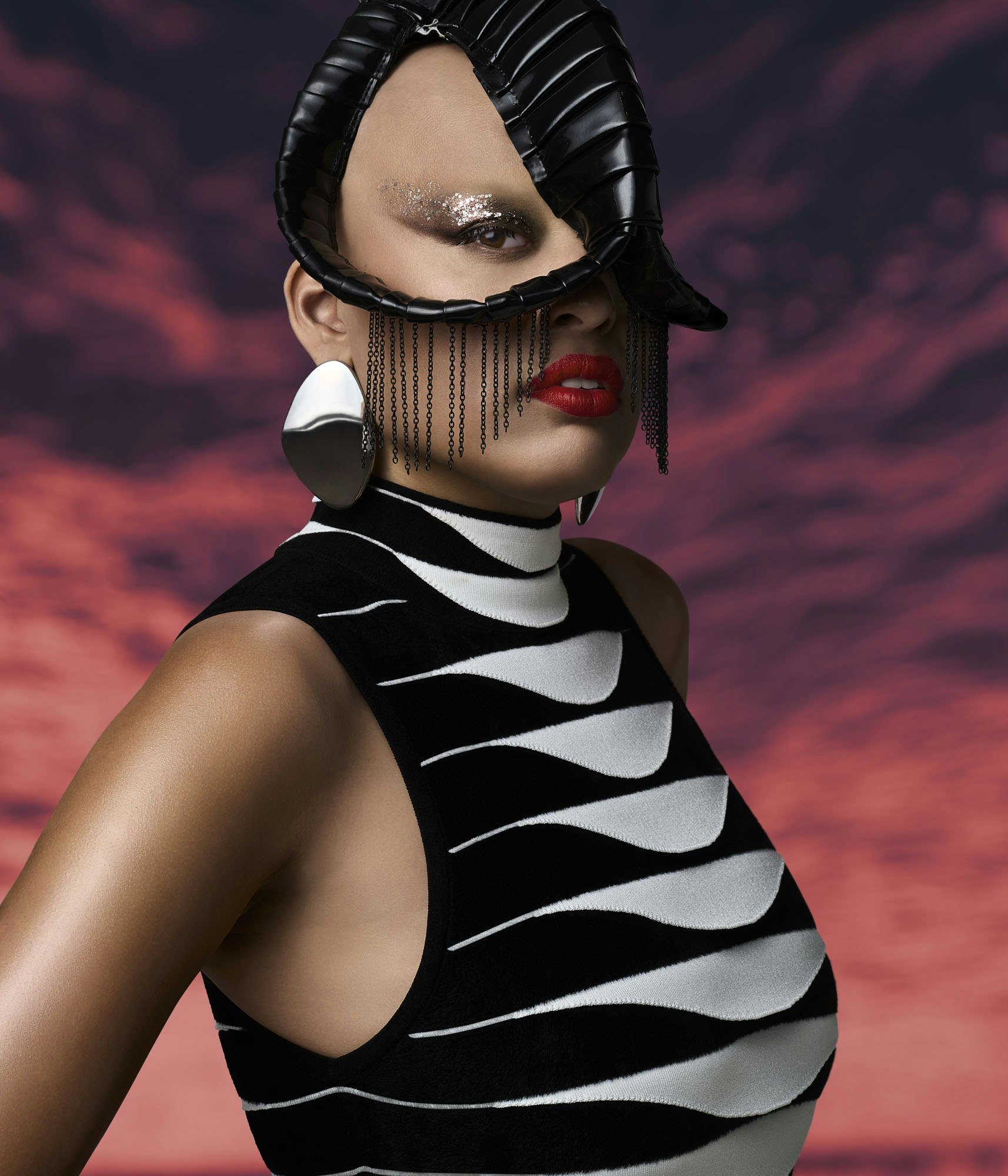 Portrait of adult woman wearing a black and white dress, dramatic makeup and sculptural headdress against a red and purple backdrop.