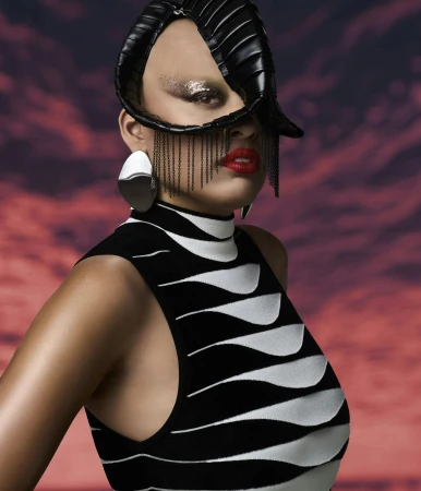 Portrait of adult woman wearing a black and white dress, dramatic makeup and sculptural headdress against a red and purple backdrop.