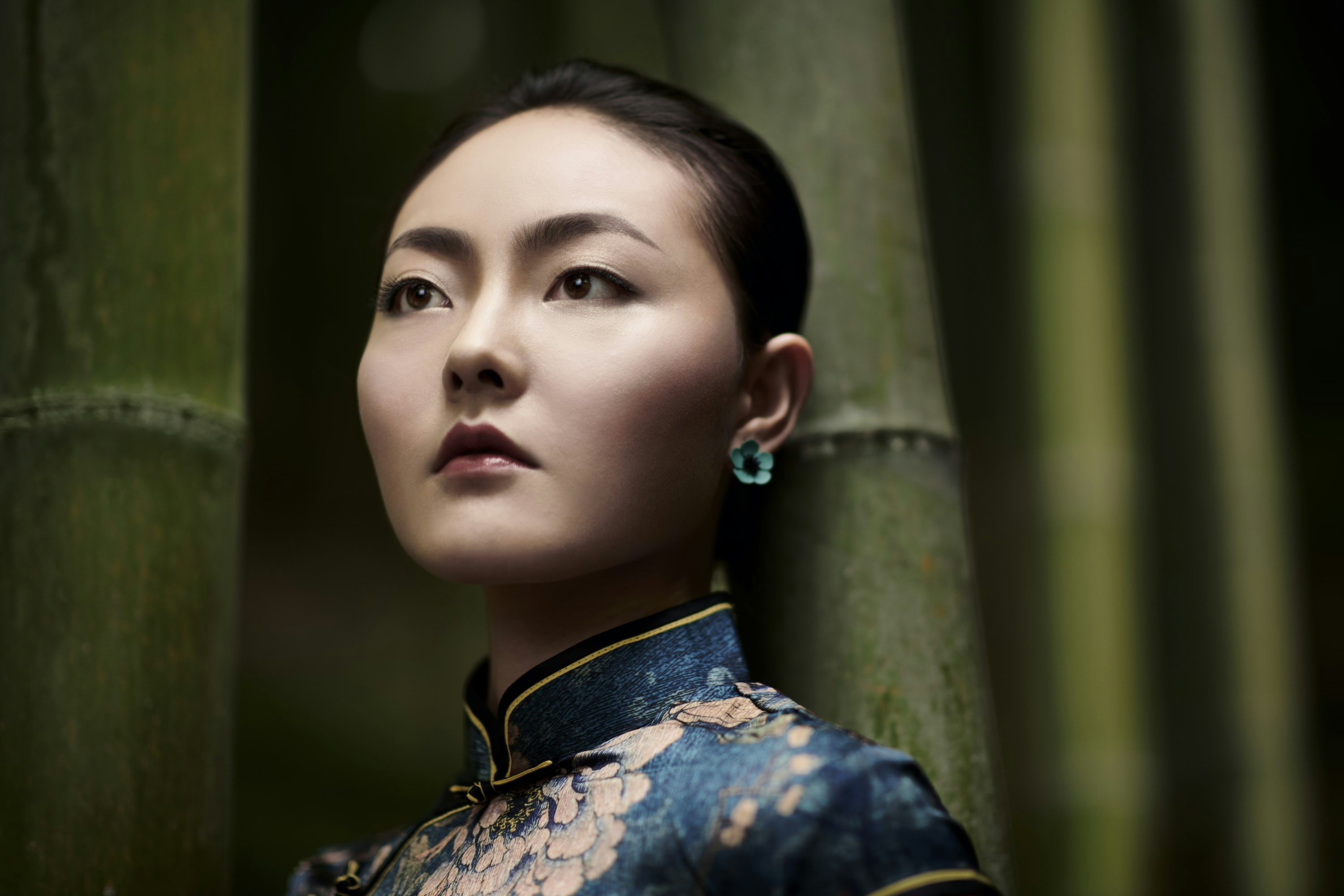 Woman with short hair in a floral dress looks up, posing in front of bamboo stalks in a close-up, eye-level outdoor portrait.