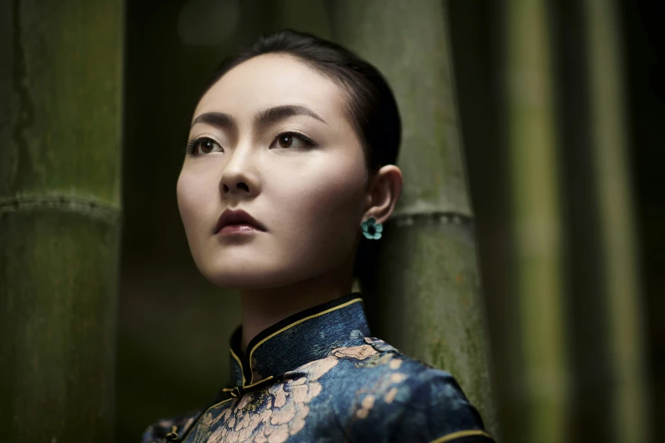 Woman with short hair in a floral dress looks up, posing in front of bamboo stalks in a close-up, eye-level outdoor portrait.