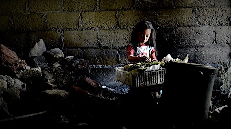 Young child sorting through items in a crate, situated against a weathered brick wall in a dimly lit, impoverished environment.