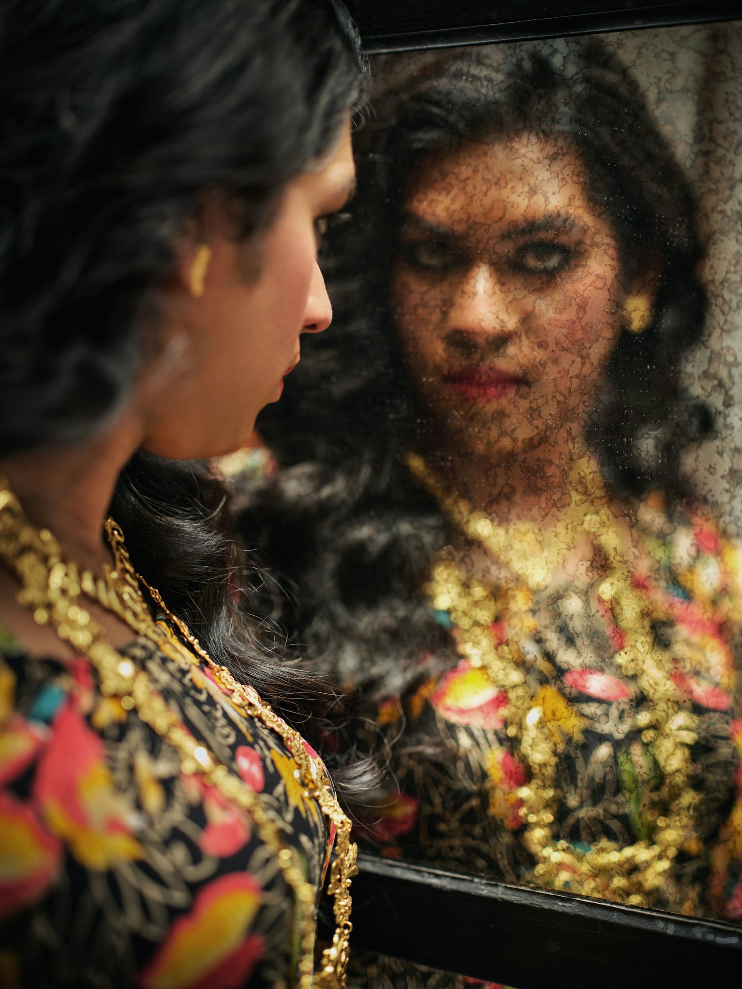 Woman with dark wavy hair wearing a patterned dress and gold jewelry looking into an antique mirror with mottled glass