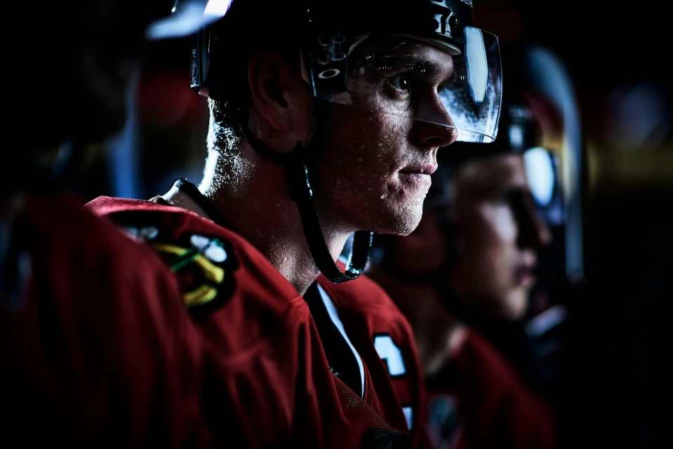 Close-up of a hockey player in a helmet and uniform, looking to the side with a determined expression, on the bench with other players.