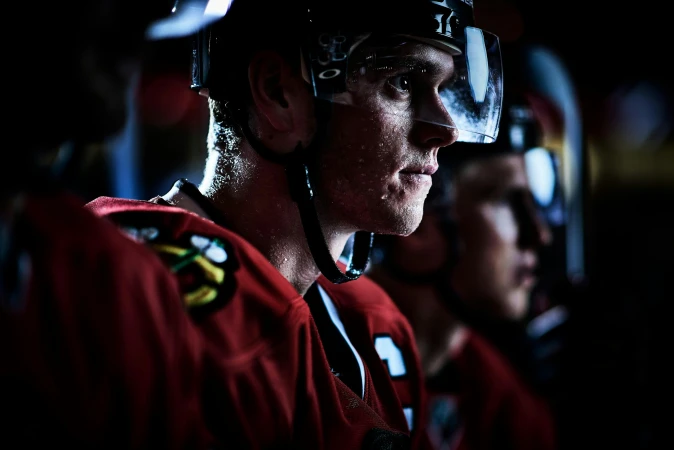 Close-up of a hockey player in a helmet and uniform, looking to the side with a determined expression, on the bench with other players.