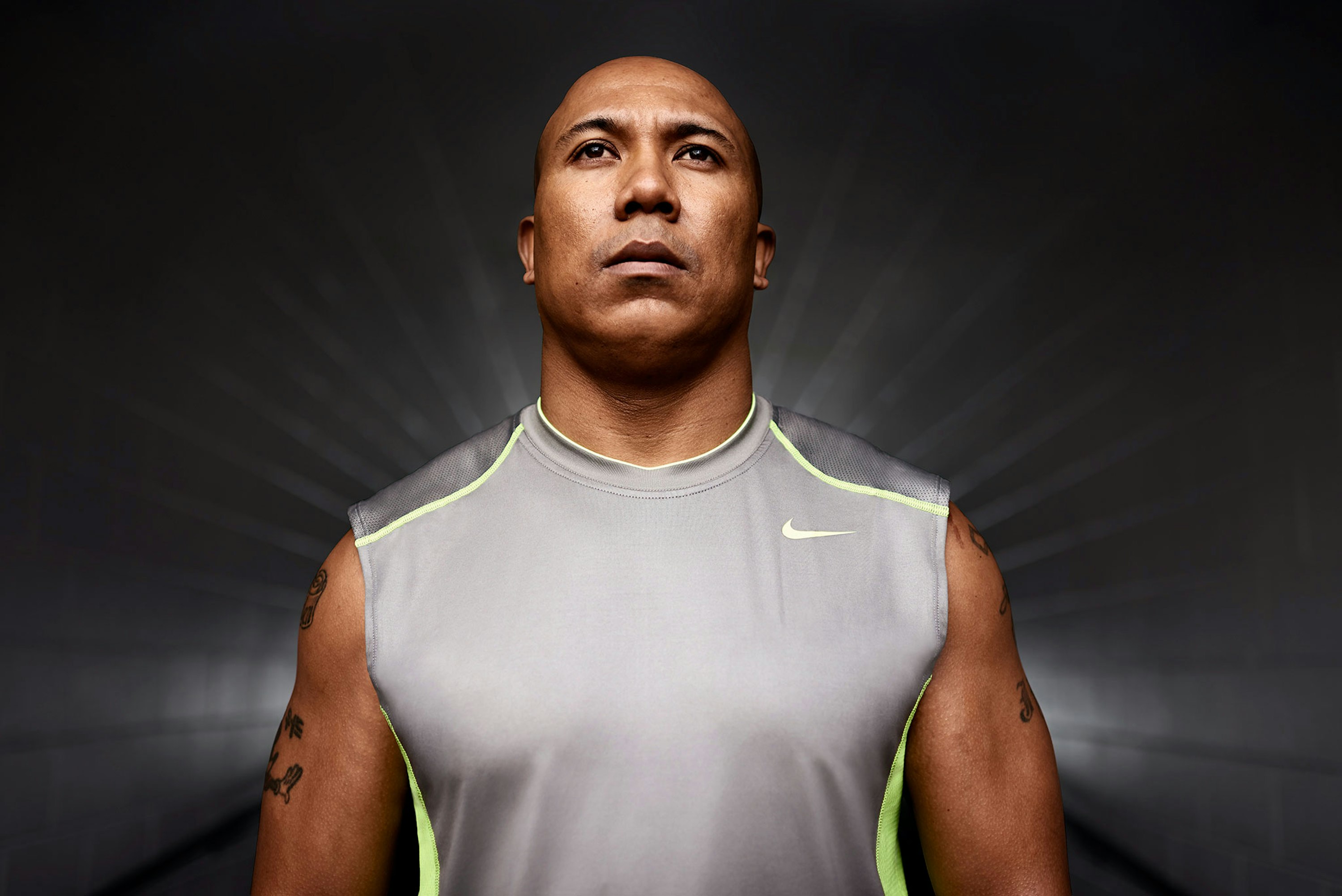 Portrait of a muscular adult man wearing a grey Nike sleeveless shirt, looking upward against a grey background with bright, diffused lighting.