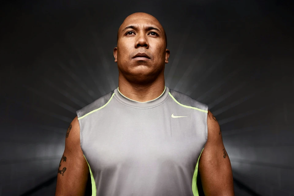 Portrait of a muscular adult man wearing a grey Nike sleeveless shirt, looking upward against a grey background with bright, diffused lighting.