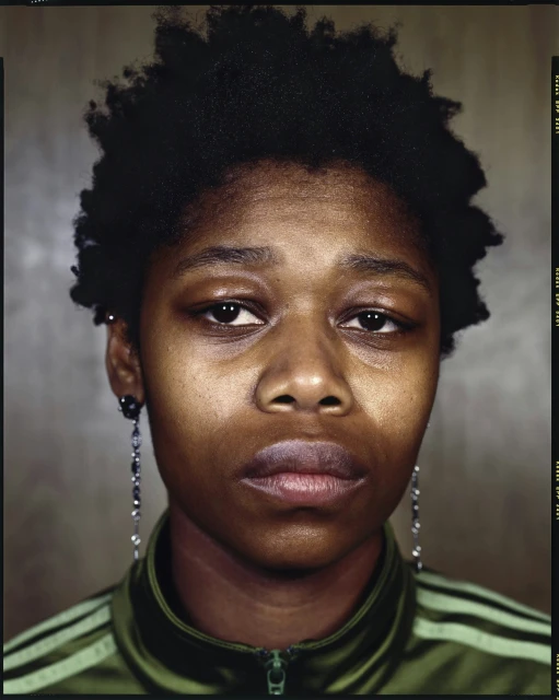 Close-up portrait of an African-American woman with an olive green tracksuit, featuring dark, cropped afro hair and dangling earrings.
