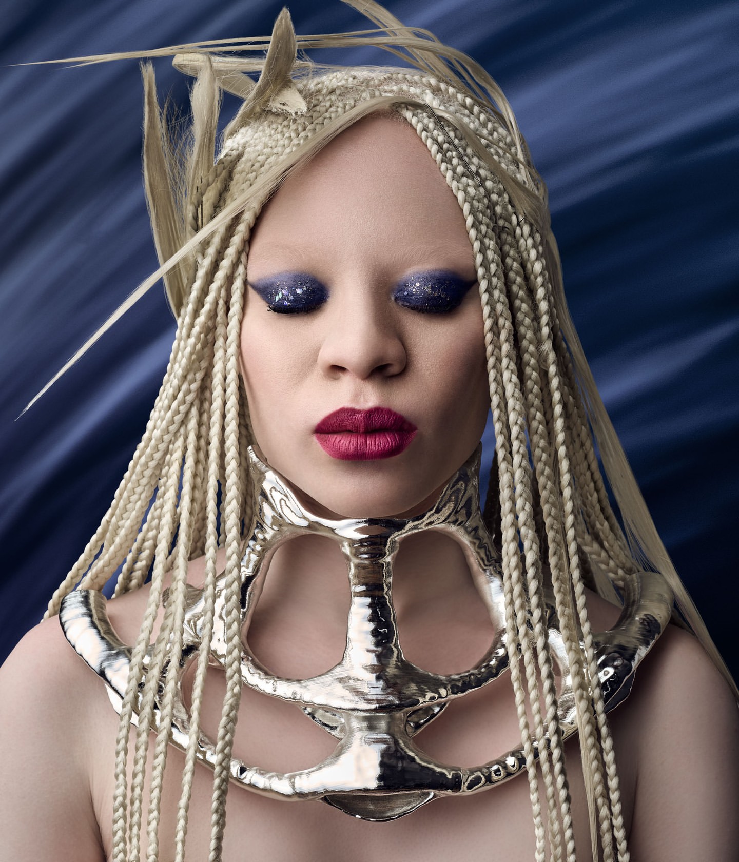 Close-up portrait of albino woman with braided hair, statement necklace, and dramatic makeup against a dark blue background.