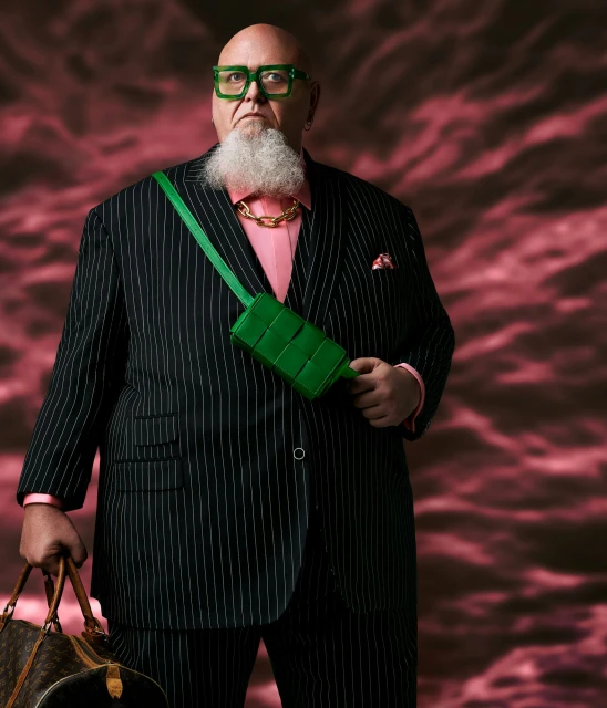 Stylish plus-size male model in pinstripe suit, green glasses, and accessories posing against a textured red backdrop in a studio shot.