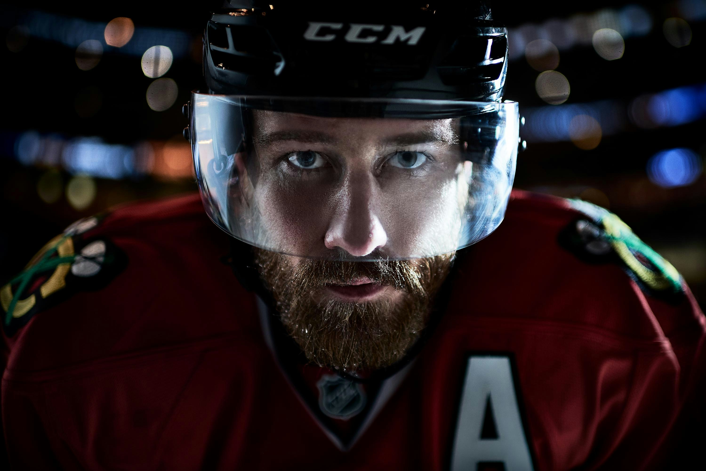 Close-up of a bearded male hockey player in a black helmet with a visor, wearing a red and black jersey, looking intensely at the camera.