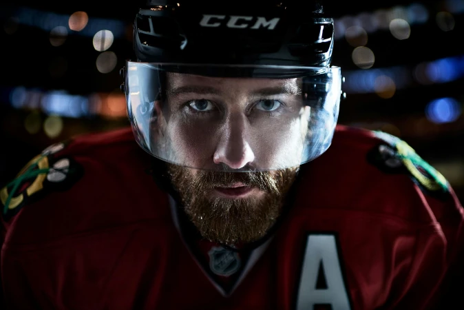 Close-up of a bearded male hockey player in a black helmet with a visor, wearing a red and black jersey, looking intensely at the camera.