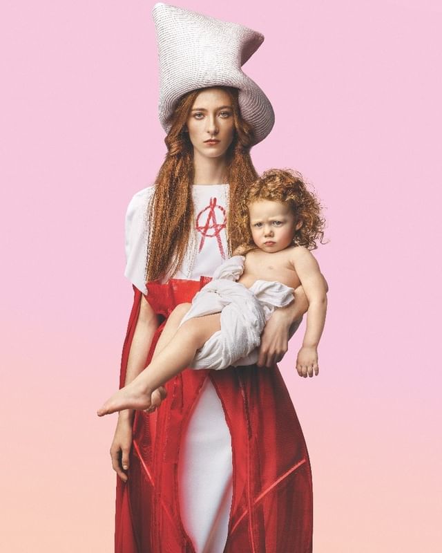 Woman and child in striking studio portrait featuring artistic styling and unique headwear against a soft pink gradient background.