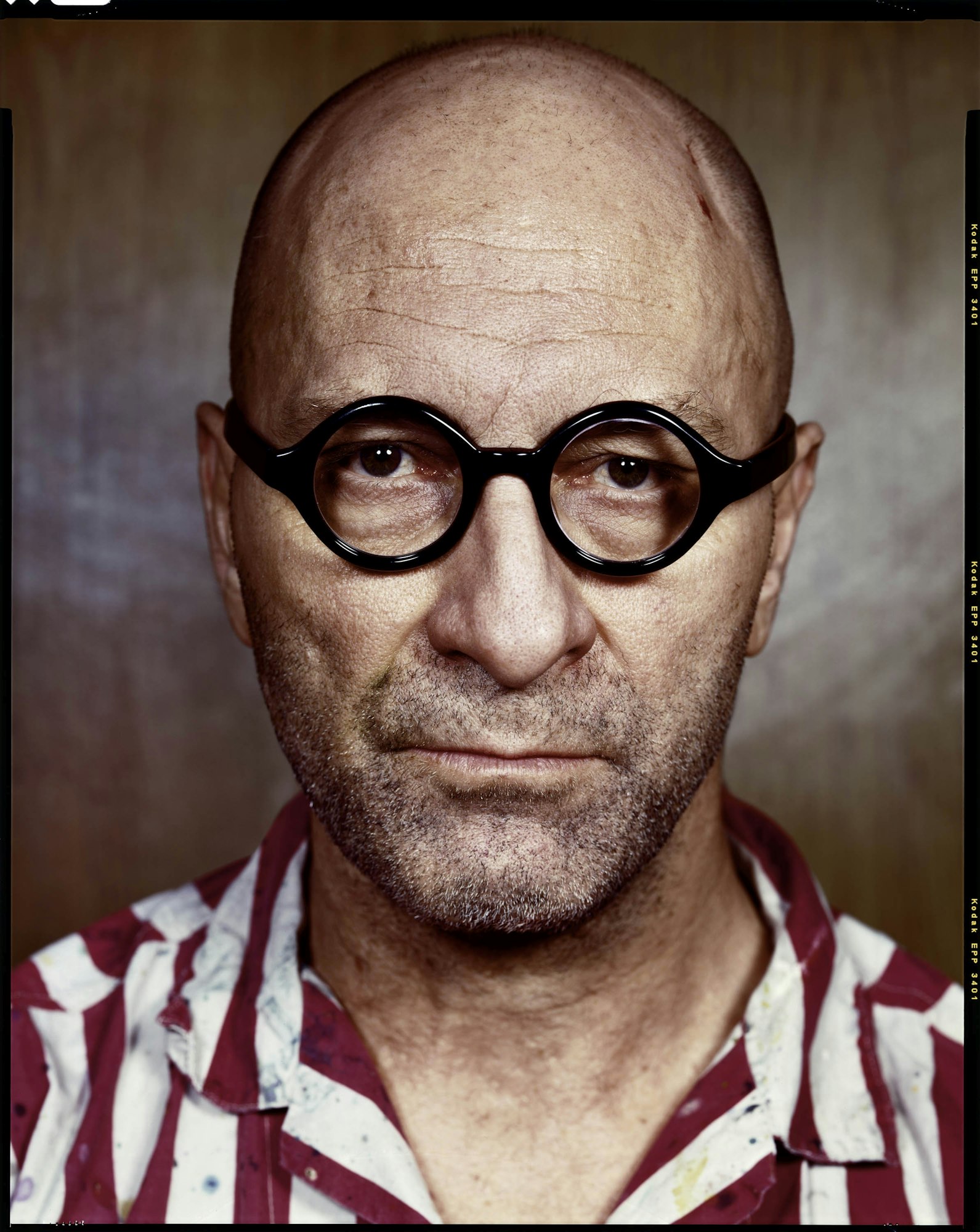 Portrait of a bald, middle-aged man with glasses wearing a red and white striped shirt against a neutral brown background.