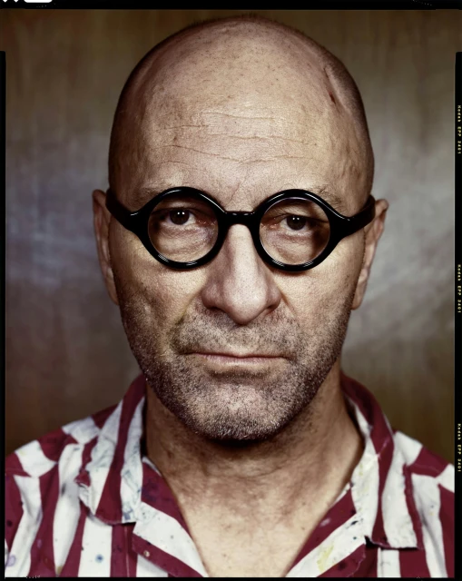 Portrait of a bald, middle-aged man with glasses wearing a red and white striped shirt against a neutral brown background.