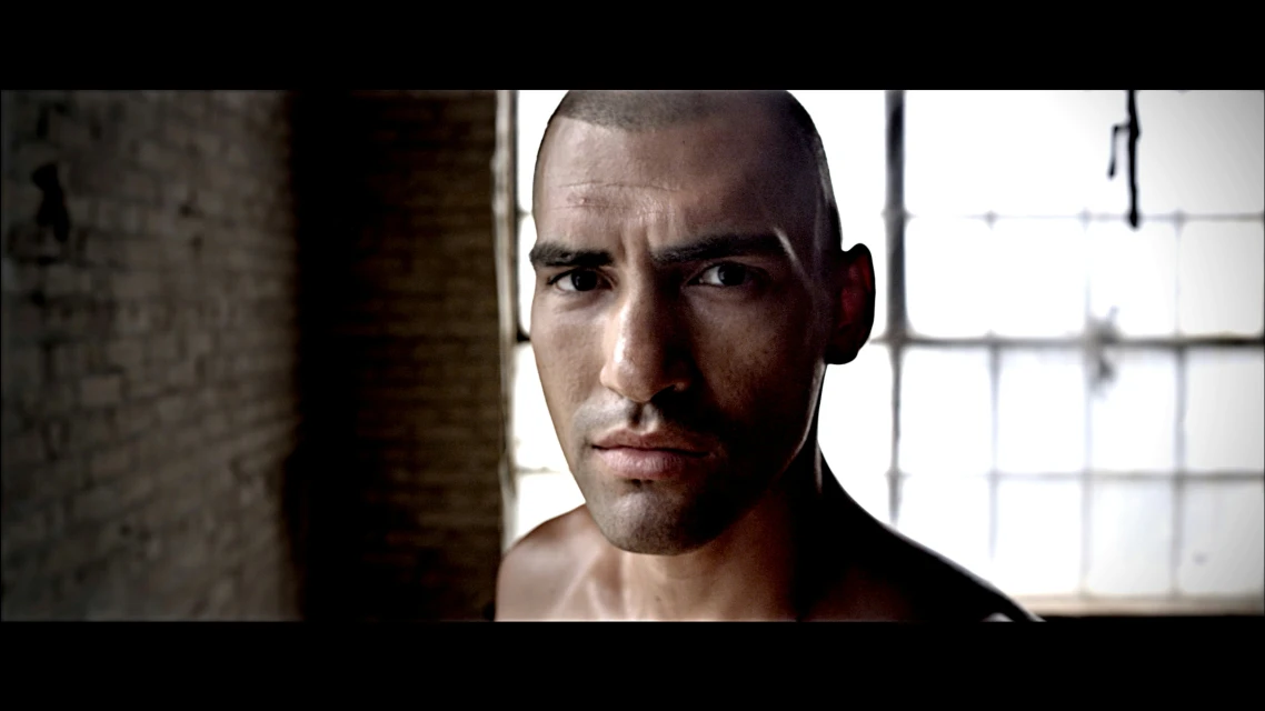 Close-up of a dark-skinned adult male looking directly at the camera in front of a blurred background of brick and a window.