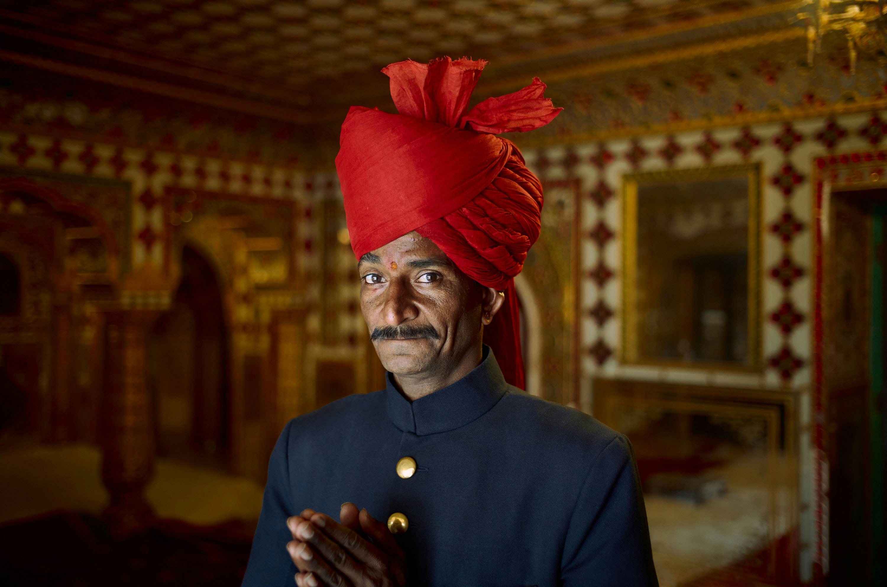 Portrait of an Indian man in a traditional turban and formal attire, standing indoors with ornately decorated walls in the background.