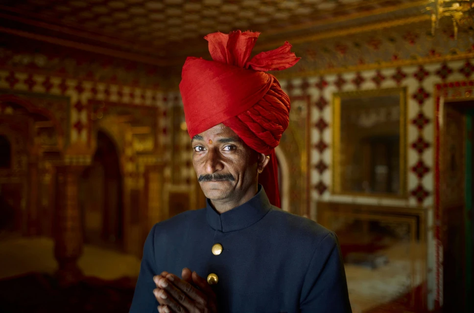 Portrait of an Indian man in a traditional turban and formal attire, standing indoors with ornately decorated walls in the background.