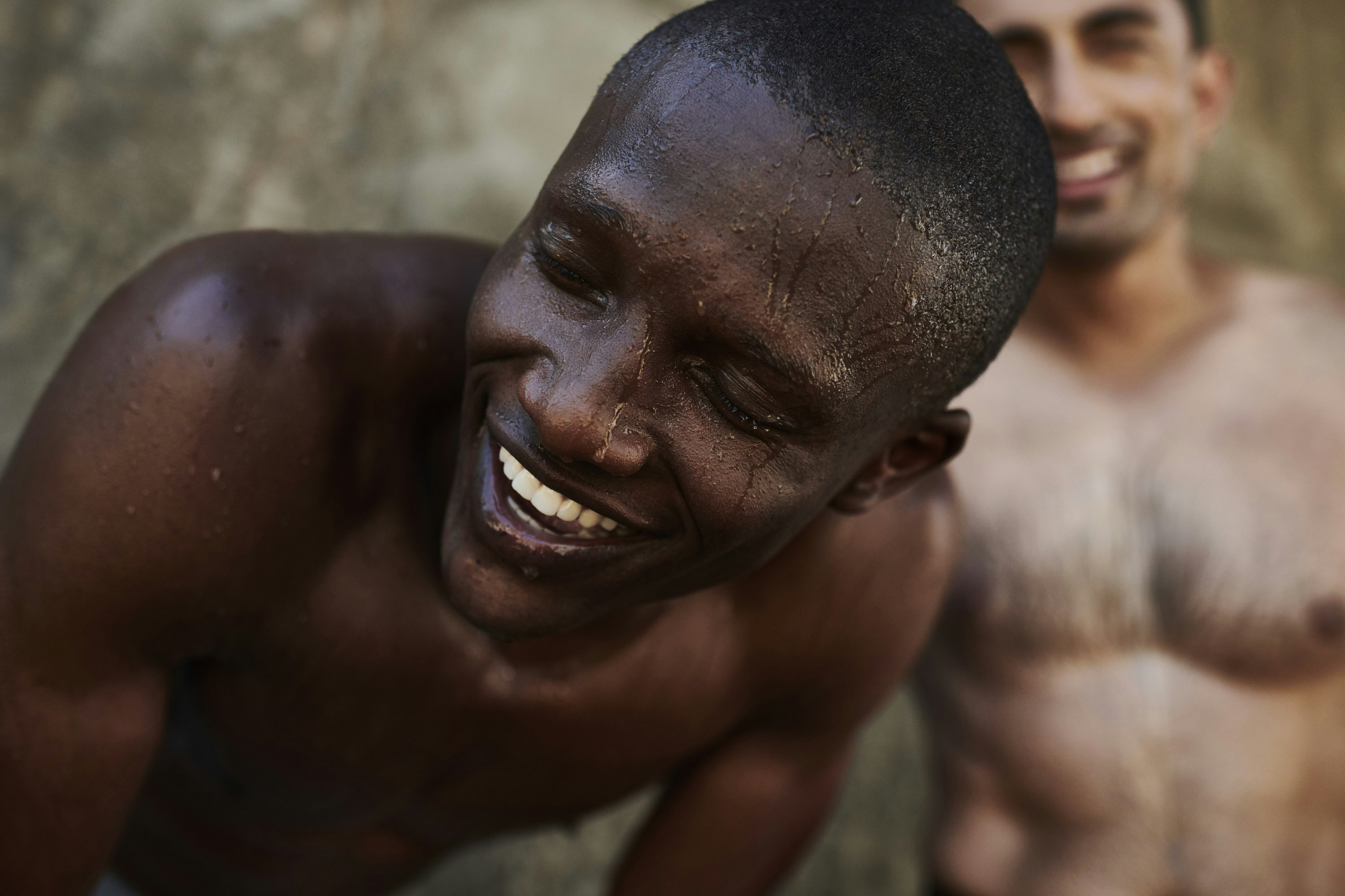 A dark-skinned man with water droplets on his face laughs, with a blurred man smiling in the background, captured in a close-up shot.