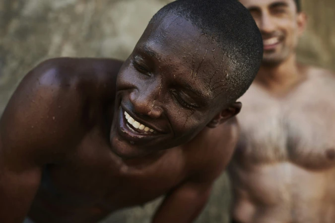 A dark-skinned man with water droplets on his face laughs, with a blurred man smiling in the background, captured in a close-up shot.