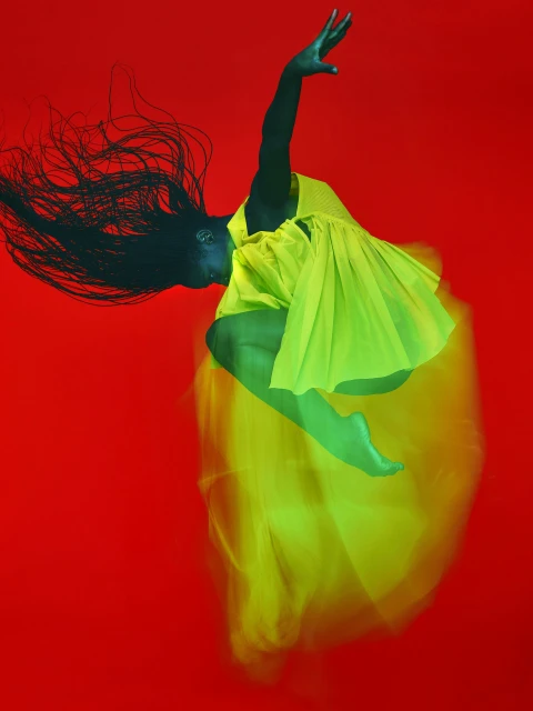 Dynamic shot of an African American woman in mid-air, wearing a bright yellow dress, against a vivid red background in a studio setting.