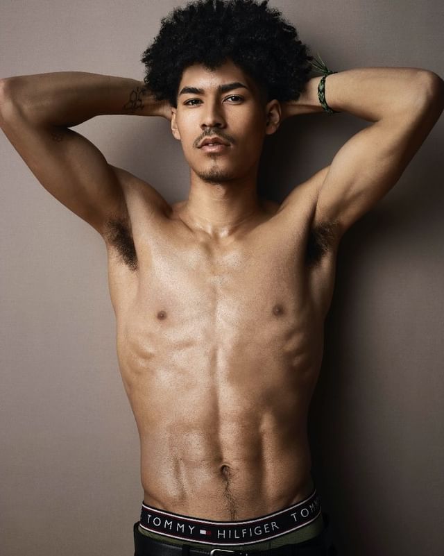 Shirtless man with an afro poses against a solid background, showcasing his physique and wearing a Tommy Hilfiger branded waistband.