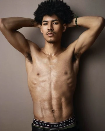 Shirtless man with an afro poses against a solid background, showcasing his physique and wearing a Tommy Hilfiger branded waistband.