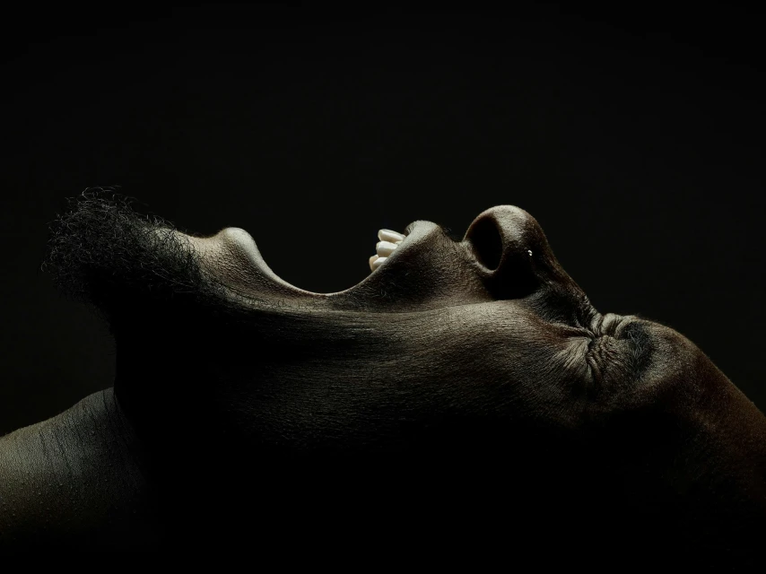 Close-up studio portrait of a dark-skinned adult male with open mouth, lit from below against a dark background. Moody, side view.