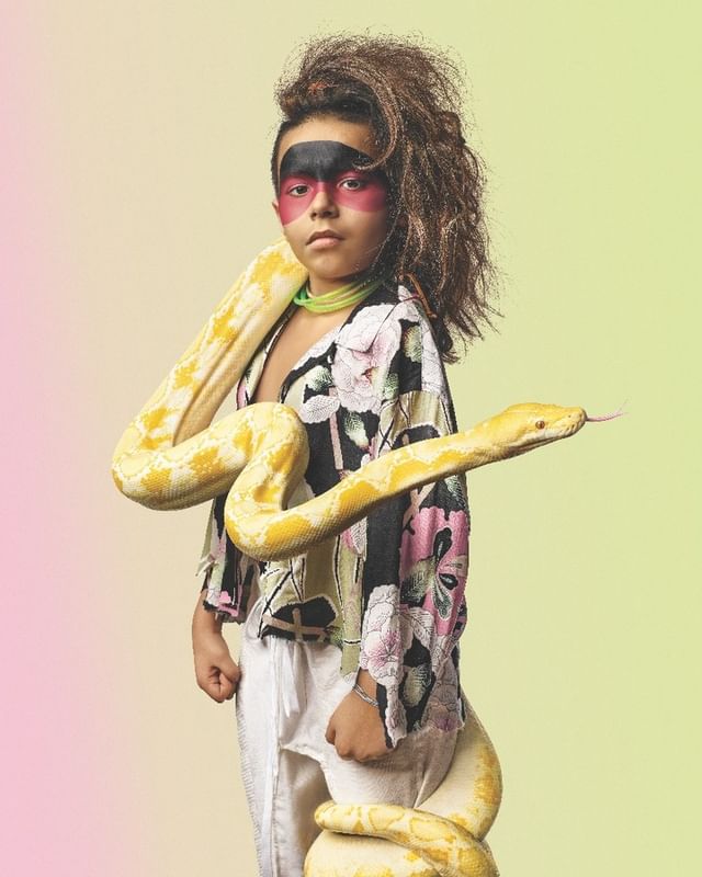 Young child with face paint posing with a large yellow snake draped around them, wearing a floral patterned shirt and white pants.