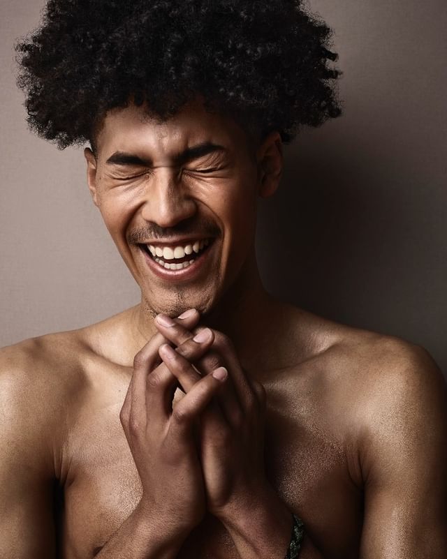 Portrait of a laughing man with dark curly hair and clasped hands against a neutral studio background