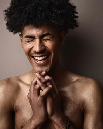 Portrait of a laughing man with dark curly hair and clasped hands against a neutral studio background