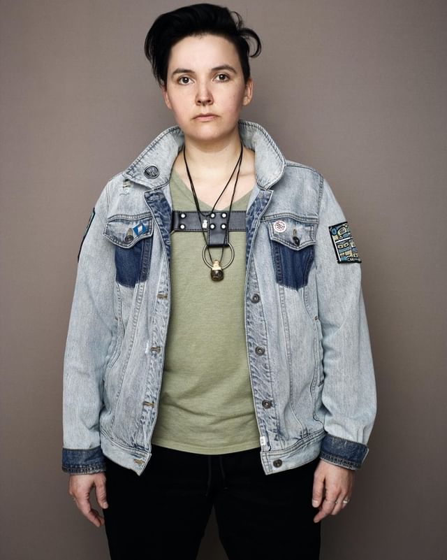 Portrait of a person with short dark hair wearing a light blue denim jacket and black pants against a neutral grey background.