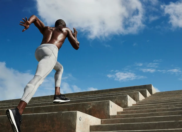 Athletic Black man running up concrete outdoor stairs against a bright blue sky with scattered white clouds.